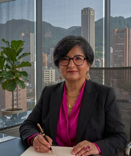 Lorena Motta, autora colombiana, sentada en una oficina con vistas a la ciudad de fondo. Viste un blazer negro sobre una blusa fucsia, lleva gafas, y escribe en un cuaderno sobre su escritorio.